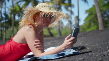 Hawaii adasındaki Pasifik Okyanusu kıyısındaki volkanik kumlu Egzotik Black Beach 'teki Mobile Phone ekranına bakan rahat genç kadın. Arka planda yeşil palmiyeli, yavaşça gülümseyen kadın.