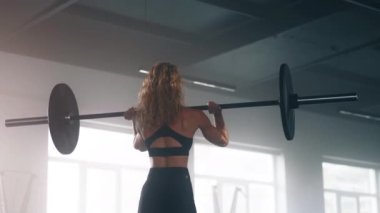 Portrait of young, active bodybuilder exercising by using a barbell. Back view of curly-haired woman lifting heavy weights on straight arms above her head. High quality 4k footage