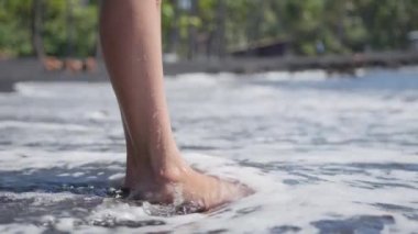 Slim female legs and feet standing at sea water waves on exotic black sand beach. Woman at seaside surf with white foamy waves splashing with foam in slow motion. Girl after bathing in ocean on shore
