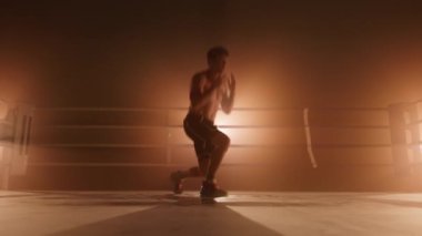 Portrait of a man with shirtless torso jumping and doing lunges to train and strengthen his leg muscles. Close-up of a squatting champion with dramatic light in the background. High quality 4k footage