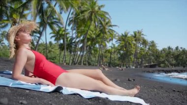Woman sitting on tropical black sand beach. Summer travel to paradise Hawaii island. Sexy girl in red swimsuit tanning at ocean with green palm trees in beautiful wild landscape.Exotic summer vacation