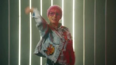 Cheerful stylish girl in pink beanie hat and pink sunglasses actively dancing, smiling in smoke and neon lights background. Actively waving hands around performing freestyle hip hot club dance moves