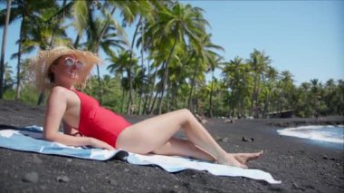 Hot young girl taking sunbath on beautiful coast summer weekend. Elegant lady wearing wide brimmed straw hat lying on black sand volcanic shore enjoying sunlight. Sexy woman relaxing in red swimsuit