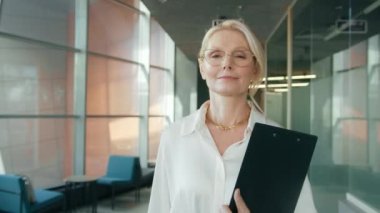 Close up portrait mature smart confident woman 50s in eyeglasses walking in corridor. Serious business woman in business center hallway. Portrait businesswoman in office corridor looking at camera 4K