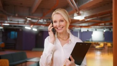 Closeup positive mature business woman 50s talking by smartphone in office interior. Smiling businesswoman calling mobile phone in corridor. Portrait of friendly CEO walking in business center hallway