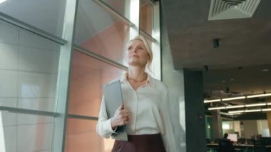 Portrait of focused businesswoman walking on meeting in office corridor. Stylish mature confident woman 50s walking in corridor. Serious business woman going with folder in business center hallway 4K
