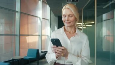 Beautiful mature woman 50s walking, using smartphone in business office hallway with daylight coming through large windows. Smiling beautiful female using mobile phone. Close-up dolly cinematic shot