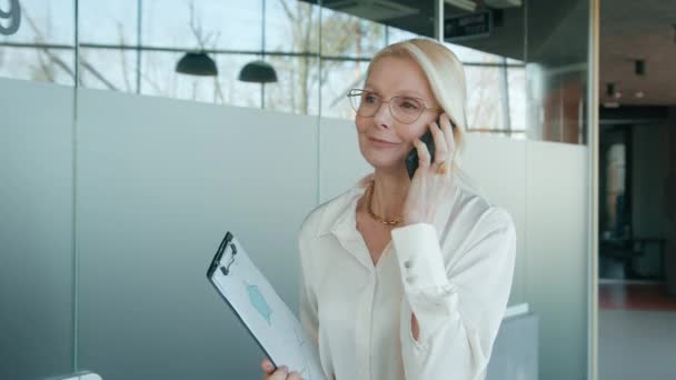 在办公室里戴着眼镜 用智能手机交谈 打扮成一个成熟的女商人 在走廊里笑着打手机的女商人友好的首席执行官走在商业走廊上的画像 — 图库视频影像