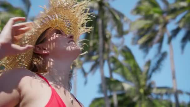 ストローハットに輝くブロンドの女性の肖像画 中年の女性の周りの柔らかいぼやけた緑のヤシの木と青空の背景に立っている パラダイス島での夏の休暇 ハワイアメリカ旅行 — ストック動画