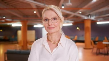 Happy mature adult global corporation manager smiling face with blonde hair in classy hairstyle, wearing glasses looking at camera, posing indoors in modern sunny modern loft office.Head shot portrait