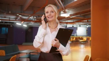 Smiling businesswoman calling mobile phone in corridor. Portrait of friendly CEO walking in business center hallway. Closeup positive mature business woman 50s talking by smartphone in office interior