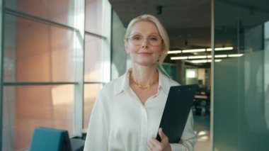 Confident business woman 40s 50s in stylish eyewear walking in business office. Independent mature female CEO executive enjoying successful corporate career. 4k footage slow motion modern loft office