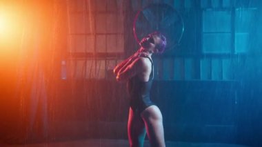 Slim young woman dancing erotic moves inside garage building or urban futuristic cyber punk studio under pouring rain in colorful spotlights. Modern professional dancer in orange blue light slow mo