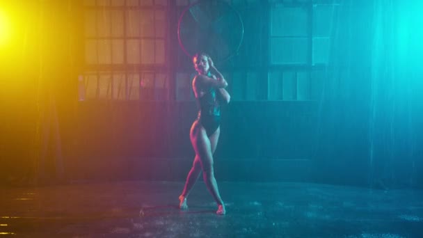 熱いダンサーは雨のガレージの背景の下の湿った床の上に座って 性的に作るエロティックなショーティールオレンジのスポットライトを移動します 誘惑女ショーセクシーボディパフォーマンスストリップサイバーパンク都市ステージ — ストック動画