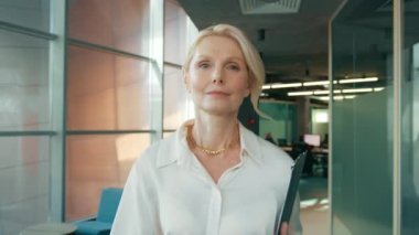 İş kadını CEO 'nun portresi ofis koridorunda 4K kamerasına bakıyor. Yetişkin, kendine güvenen 50 'li yaşlarda bir kadın koridorda yürüyor. Ciddi bir iş kadını iş merkezi koridorunda panoyla geziyor.