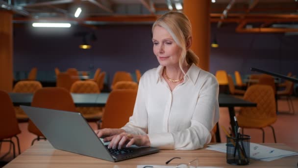 年长的职业成熟的商业女性50岁 使用笔记本电脑坐在工作场所的办公桌前 在现代的开放式办公室里 快乐的高级职员灰白头发的女行政人员在个人电脑上打字 — 图库视频影像