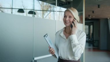 Gülümseyen olgun bir kadın resmi gömlek giyiyor iş ortaklarıyla pazarlık yapıyor cep telefonuyla sözleşme yapıyor, elinde sunum panosuyla şirket ofisindeki sunum panosuyla yürüyor, yavaş çekim.
