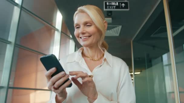 用手机微笑美丽的女性 特写娃娃电影拍摄 漂亮的50多岁的成熟女人走着 在有大窗的商业办公室走廊里用智能手机 — 图库视频影像