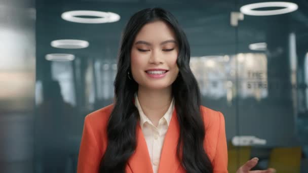 会議で若い女性を自信を持って会話を呼び出します ポートレートゴージャスダークヘア若いアジアの女性は オレンジ色のブレザーを身に着けている中間近代的なオープンスペースビジネスオフィスに立っている間魅力的に微笑む — ストック動画