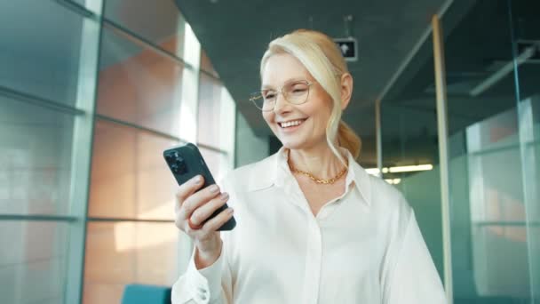 スマートフォンを持ちながらオフィスを歩く熟女の笑顔 携帯電話のモバイルアプリを使用して50歳のマネージャーCeo ビジネス企業のためのデジタル技術アプリケーションとソリューション スローモーション — ストック動画
