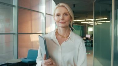 Kendine güvenen iş kadını 40 'lı yaşlarda, iş yerinin koridorunda yürüyor. Gülümseyen, olgun kadın CEO, başarılı şirket kariyerinin tadını çıkarıyor. 4k video yavaş çekim modern çatı katı ofisi