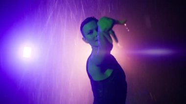 Dalış kıyafetli genç zarif bayan dansçının sinematik yakın plan çekimleri, arka planda dramatik yağmur ve ışıklandırma ile şehir garajında erotik baştan çıkarıcı koreografi sergiliyor.