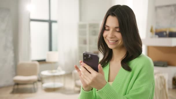 現代のアパートの背景にスマートフォンを使って屋内に立つ美しい女性 携帯電話を使って美しい女性を笑顔 人々の技術は映画的なドルリースローモーションを閉じます — ストック動画