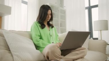 Genç esmer kadın üniversite öğrencisi dizüstü bilgisayarla öğreniyor, notlar yazıyor, webinar ya da sanal eğitim izliyor, uzaktan eğitim görüyor ve modern apartmanda uzaktan eğitim görüyor.