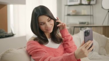 Güzellik modası sosyal medya blogcusu akıllı telefondan ön kameraya bakarak selfie çekiyor. Mutlu genç esmer kadın cep telefonu kamerası ve güzel moda maskesiyle uygulama kullanıyor. Kanepeye otur.