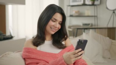 Kanepede otururken akıllı telefon kullanan neşeli genç bir kadın. Modern bir dairede cep telefonu uygulaması kullanarak gülümseyen güzel bir kadın. Güzel kız evde cep telefonuyla sohbet ederken dinleniyor.