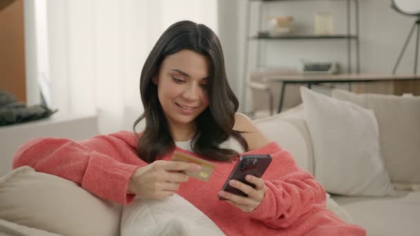 現代の家でソファに座っているスマートフォンやクレジットカードを持っている若い女性を笑顔 即座の簡単なモバイル決済を使用して幸せな女性の買い物客 オンラインストアでの顧客の購入 Eバンキングアプリ — ストック動画