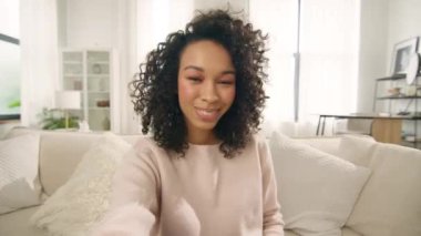 Genç, Afro-Amerikan melez, video blogcu kadın el sallıyor, webcam 'e bakıyor, hipster kız kamerayla konuşuyor video konferans görüşmesinde kanepede oturuyor. Sosyal uzaklıkla buluşan sanal sohbet arkadaşları