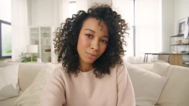 Sanal sohbet arkadaşları sosyal mesafeyle buluşuyor. Kamerayla konuşan mutlu hippi kız video konferans görüşmesinde kanepede oturur. Genç Afrikalı Amerikalı melez vlogger kadın el sallayarak web kamerasına bakıyor.