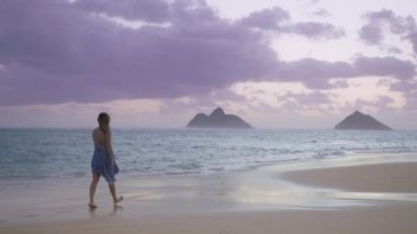 Mokulua adaları manzaralı güzel Lanikai kumlu plajında çıplak ayakla yürüyen mutlu kadın sabah gökyüzünde pembe bulutların doğuşunu izliyor. Hawaii 'ye yaz tatili için ilham verici. Kırmızı Kamera 4K.