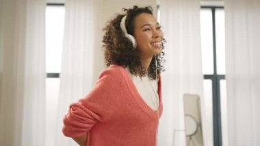İçeride müzik dinleyen kulaklıklı Afrikalı Amerikalı genç bir kadının portresi. Şirin afro saç stili şarkı söyleyen, şık tasarım dairesiyle dans eden melez bir kadın. Ağır çekimde 4K