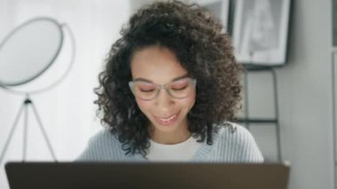 Profesyonel neşeli melez esmer kadın dizüstü bilgisayarda yazıp bitirince kapatıyor. Kıvırcık saçlı genç bir kadın önemli bir e-posta yazıyor. Modern ev oturma odası. Uzak çalışma konsepti 4K