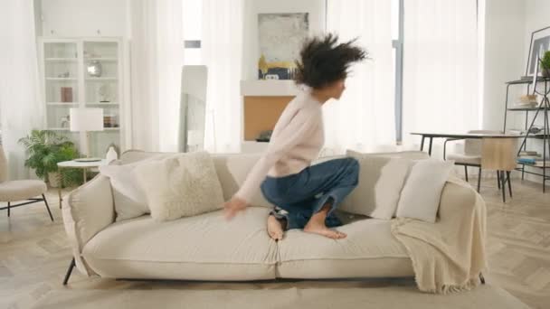 モダンなデザインのインテリア家具の広告コンセプト 笑う少女は家に一日で移動を祝う フラットを借り 居心地の良いソファの上で休息を楽しんで 喜びを感じている 幸せなアフリカ系アメリカ人女性ジャンプソフトソファ — ストック動画