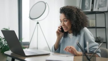 Gülümseyen siyahi iş kadını bilgisayarla telefonla konuşuyor. Ofis masasında oturuyor. Mutlu karışık ırk bayan müşterileri internet sitesi alışveriş siparişi konseptini doğrulamak için mobil arama yapıyor.