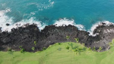 Yeşil golf sahasındaki siyah lav kayalıkları, açık mavi su dalgalarıyla şeffaf mavi okyanus manzarası. Egzotik Büyük Ada Hawaii, ABD turizminin en güzel yeşil palmiyesi.