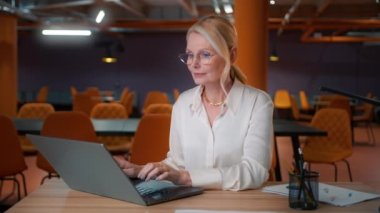 Olgun yetişkin CEO kadın profesyonel pazarlama yöneticisi dizüstü bilgisayar kullanıyor. Bilgisayar dijital veri yönetimi üzerine çalışan yorgun bayan iş kadını 4K gecesinde büyük modern şirket ofisinde oturuyor.
