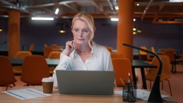 50岁的女商人在笔记本电脑上输入电子邮件 喝咖啡 带着眼镜的疲倦的女老板在现代阁楼风格的办公室工作 背景是运动的 有魅力的晚班妇女 — 图库视频影像