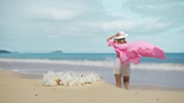 Yakın çekim tropikal beyaz orkide çiçekli kolye. Okyanusa koşan, uçan pembe elbiseli, kaygısız kadın. Zarif kıyafetli özgür neşeli kız neşe ve mutluluk içinde dinleniyor, kusursuz Oahu beyaz kumu.
