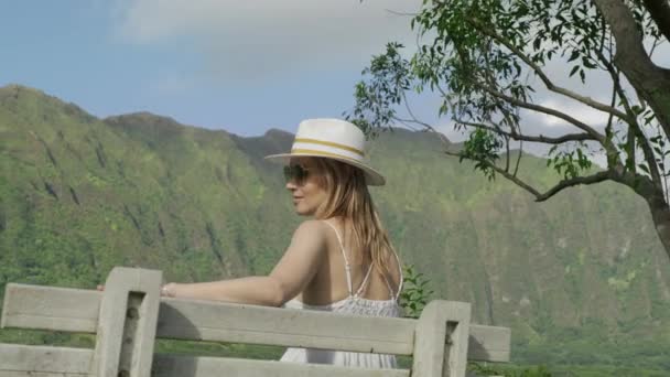 スタイリッシュな女性観光客は 動きの背景に高い緑のジャングルの山の素晴らしい景色と熱帯公園の木製のベンチに座っています カジュアルBohoスタイルのドレスと白の帽子 背景色を好転させる Red — ストック動画