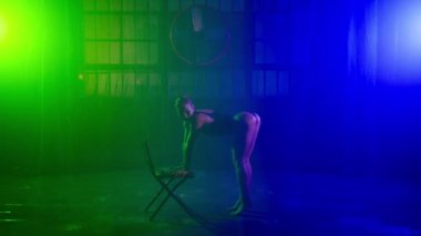 Genç seksi dansçı ıslak dans pistinde yağmur altında sandalyede gösteri yapıyor. Renkli spot ışıklarında dans eden güzel bir kadın. Esnek kız gece kulübünde sandalyede jimnastik striptiz yapıyor.