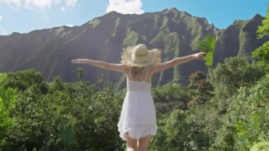 Hasır şapkalı şık bir kız ve Hawaii 'nin yüksek ormanlarındaki dağ manzaralı beyaz boho elbisesi Oahu seyahati. Neşeli, mutlu, turist bir kadın. Kocaman yeşil bir tropik yaprak ile mutlu bir şekilde el kaldırıyor.