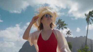 Kırmızı mayo giymiş lüks seyahat eden kadın arka planda manzaralı Hawaii manzarasında yürüyor. 30 'lu yaşlarda bir kadın kıyıda rüzgarın tadını çıkarıyor. Yaz tatili boyunca güneş şapkalı zarif bayan Kualoa çiftlik manzaralı 4K