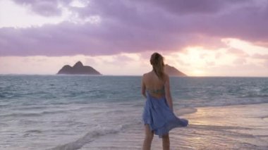 Oahu Hawaii Adası ABD 'de yaz tatilindeki kadın turist. Yavaş çekim yapan kadın, altın gün doğumunda güzel sahilde yürüyor. Güzel sallanan elbiseli kadın sinemada altın gün doğumu REC kamera görüntüsü