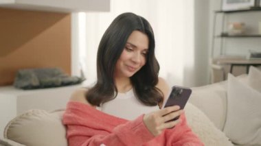Ekonomi sınıfında oturan güzel bir kadın, modern bir dairede hareketli arka planda akıllı telefon kullanıyor. Gülümseyen güzel bir kadın cep telefonu kullanıyor. Dolly sinemasını kapatın. İnsanları ve teknoloji konseptini çekin.