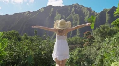 Hasır şapkalı şık bir kız ve Hawaii 'nin yüksek ormanlarındaki dağ manzaralı beyaz boho elbisesi Oahu seyahati. Neşeli, mutlu, turist bir kadın. Kocaman yeşil bir tropik yaprak ile mutlu bir şekilde el kaldırıyor.