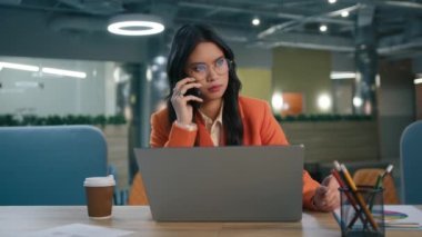 Gözlüklü karışık ırk kadın yöneticisi cep telefonu ticareti anlaşmasıyla proje detaylarını tartışıyor. Konsantre Asyalı iş kadını telefonda dizüstü bilgisayarlı ofis masası kullanarak konuşuyor.